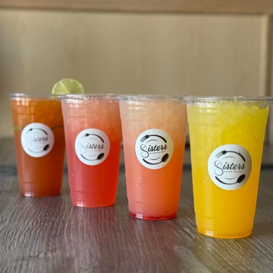 Four colorful Sisters Asian Eatery drinks, including orange, red, pink, and yellow, on a wooden surface.