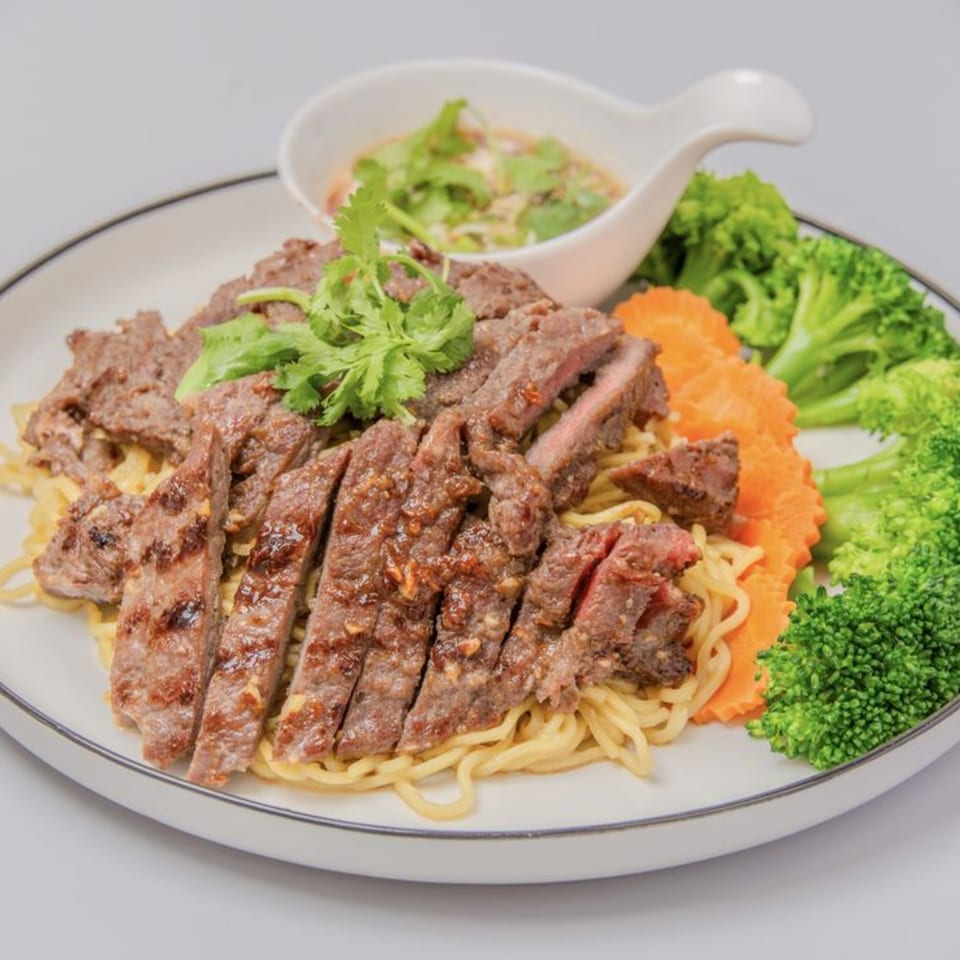 Sliced grilled beef over noodles with broccoli, carrots, and a side of dipping sauce.